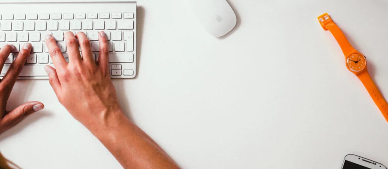 Hands typing on a keyboard with a mouse, orange watch, and smartphone on a white desk.