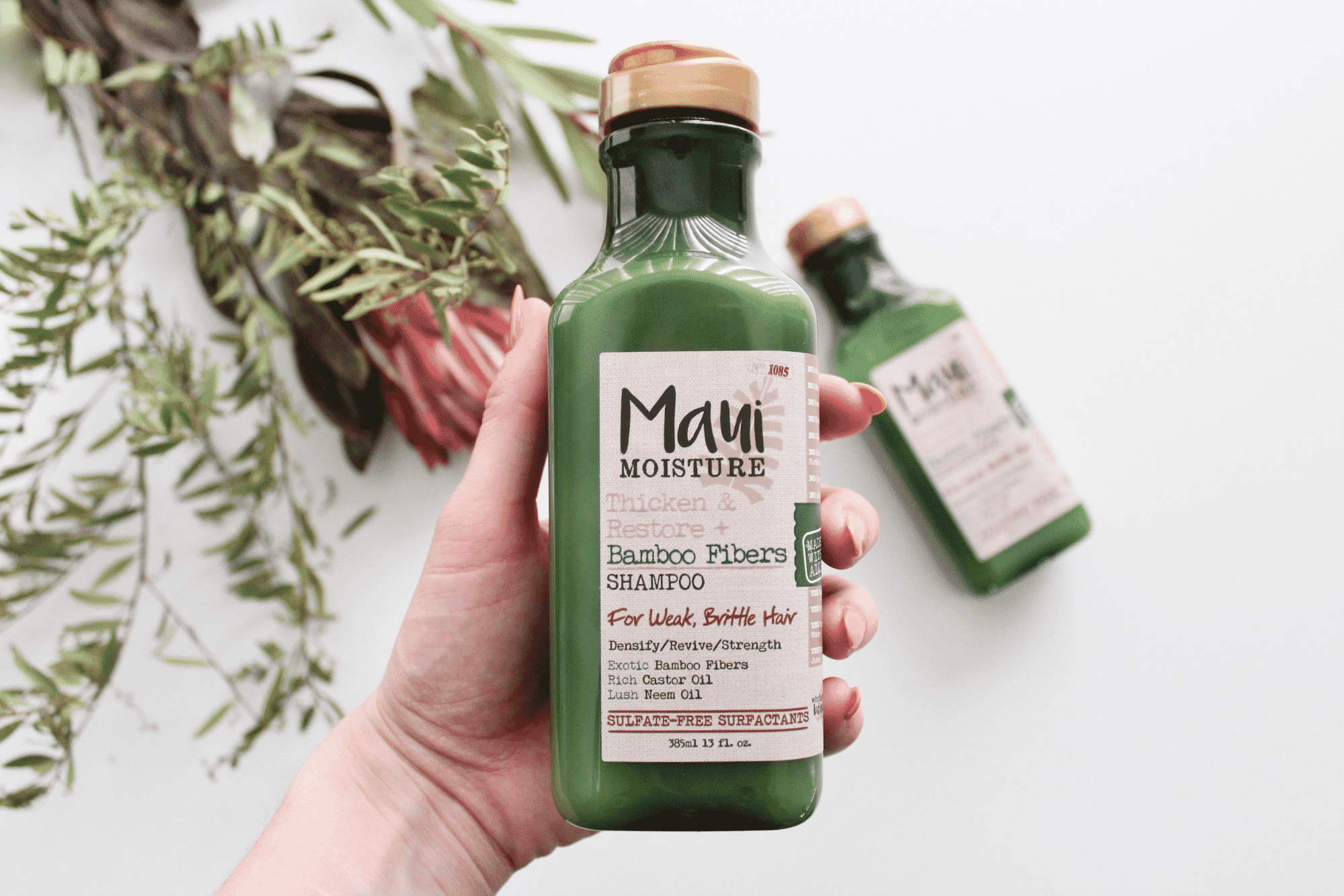 Hand holding Maui Moisture Bamboo Fibers Shampoo bottle with greenery and a second bottle in the background