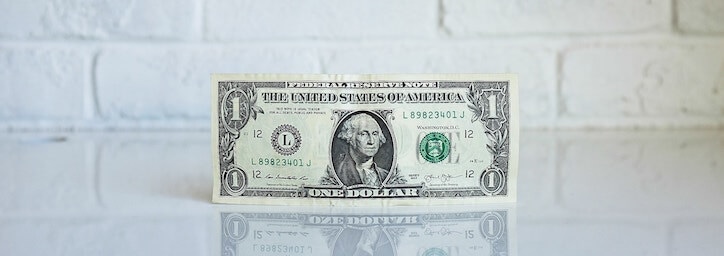 Single U.S. dollar bill standing upright on reflective surface with white brick background.