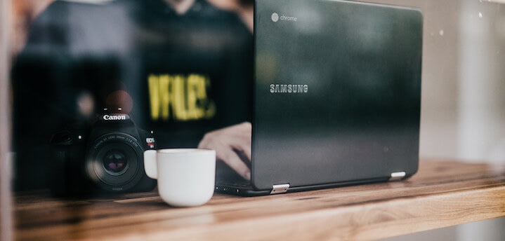 Person working on Samsung Chromebook with Canon camera and coffee mug on wooden table in a relaxed setting.