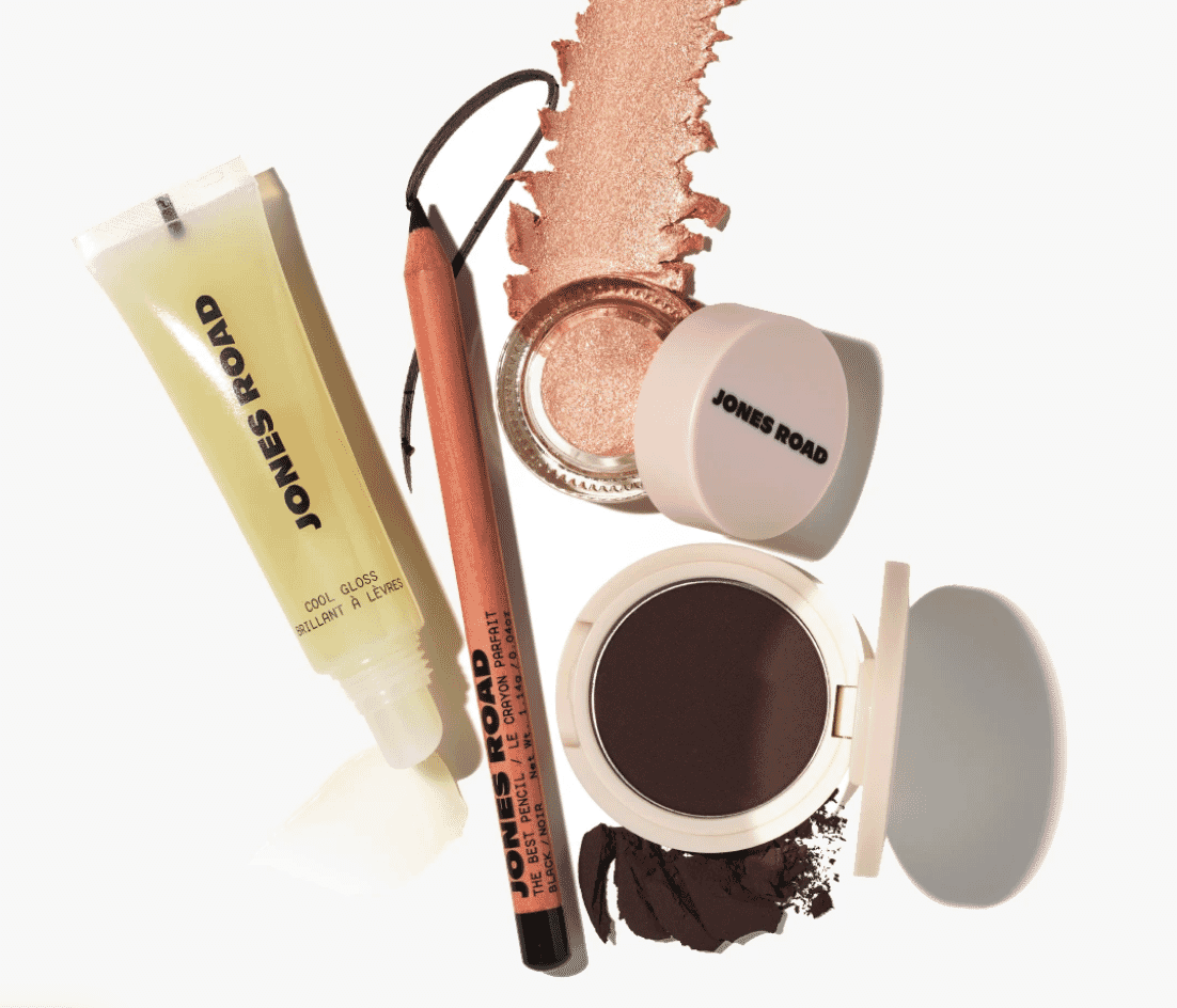 Flat lay of Jones Road beauty products, including lip gloss, eye pencil, shimmering powder, and brown eyeshadow compact.
